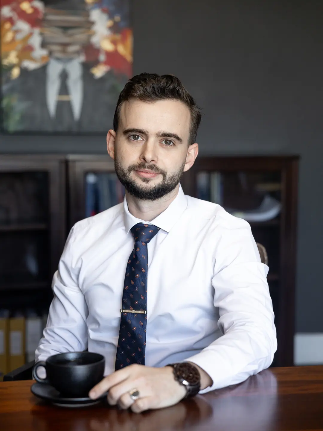 Attorney dressed in collar and tie seated at a table in a professional work environment with a cup of coffee